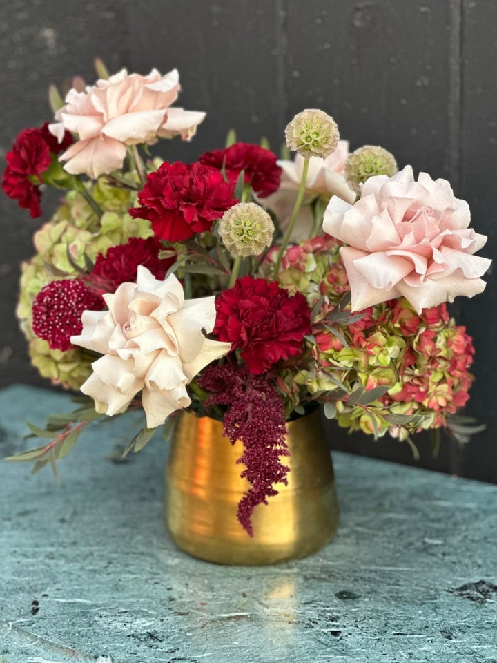 Bouquet cranberry and burgundy flowers in gold vase sitting on a textured table in front of a black wall. 