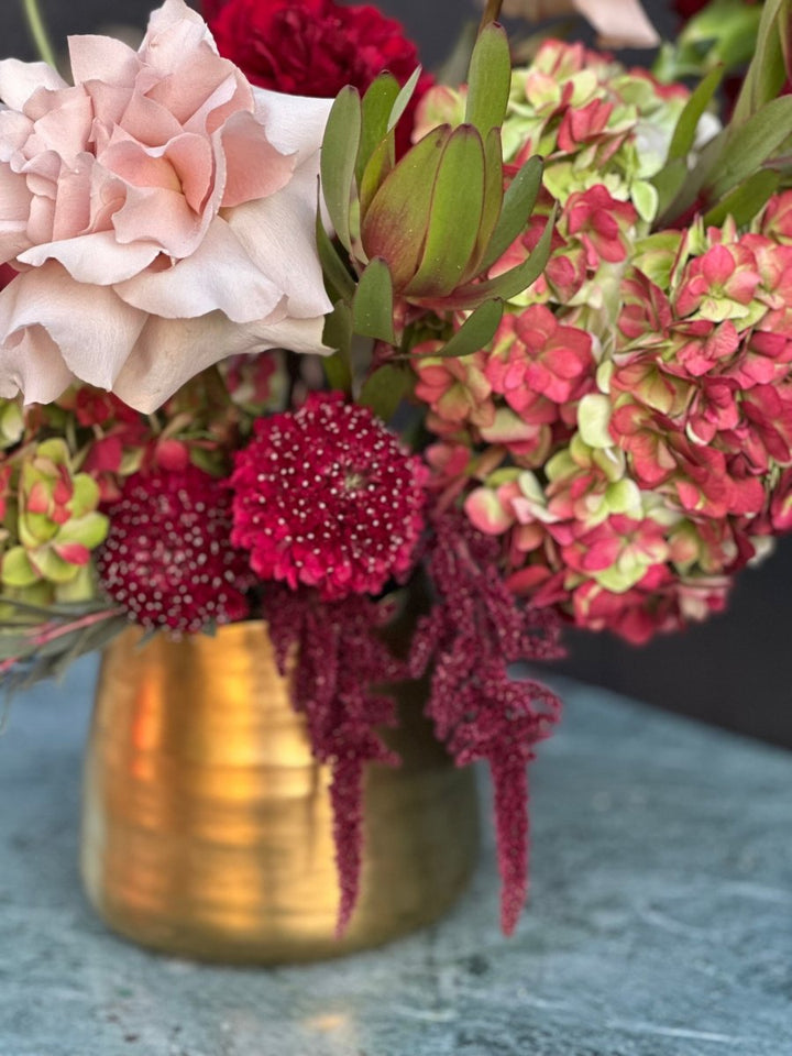 close up of cranberry colored scabiosa and quicksand rose next to hydrangea in gold vase. 