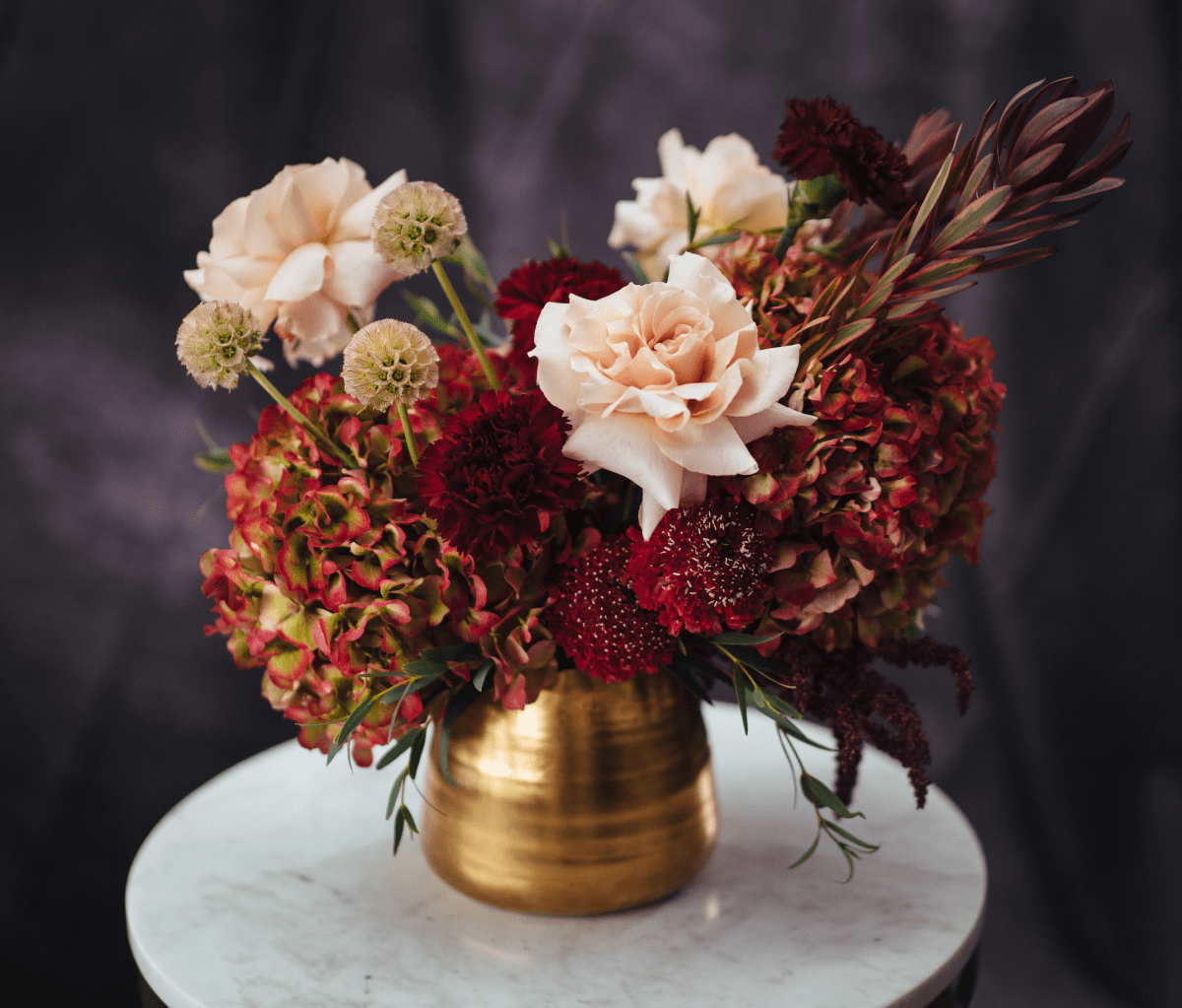Autumn floral arrangement with peach roses, burgundy dahlias, and bronze foliage in gold brass vase on marble table