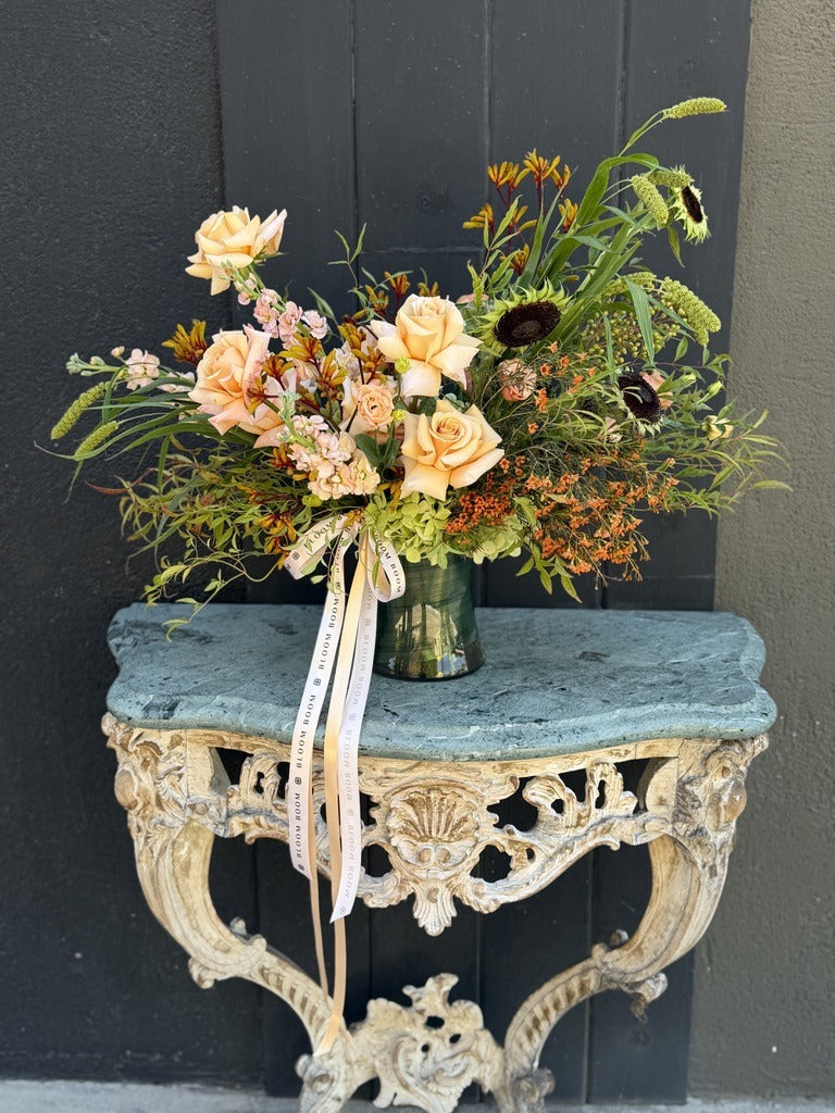 Autumn Safari fall arrangement of peach roses, blush stock, kangaroo paw, sunflowers and orange waxflower in a green glass vase with cream ribbons