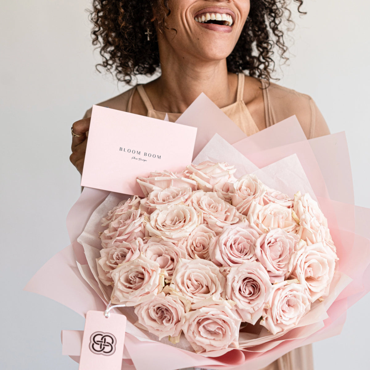 Smiling woman holding luxury bouquet of 25 blush pink roses wrapped in soft pink paper with Bloom Boom branding