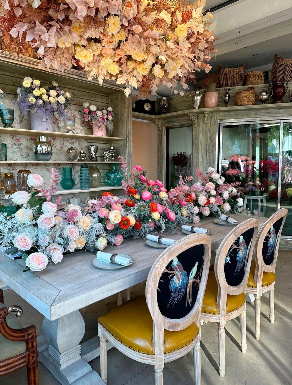 Long dining table set with pastel floral centerpieces and hand-painted bird chairs inside the Bloom Boom event space