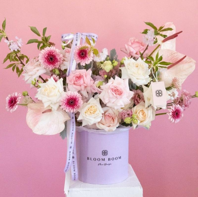 Lavender hat box filled with pink roses, white blooms, and burgundy mums on pink background with Bloom Boom ribbon