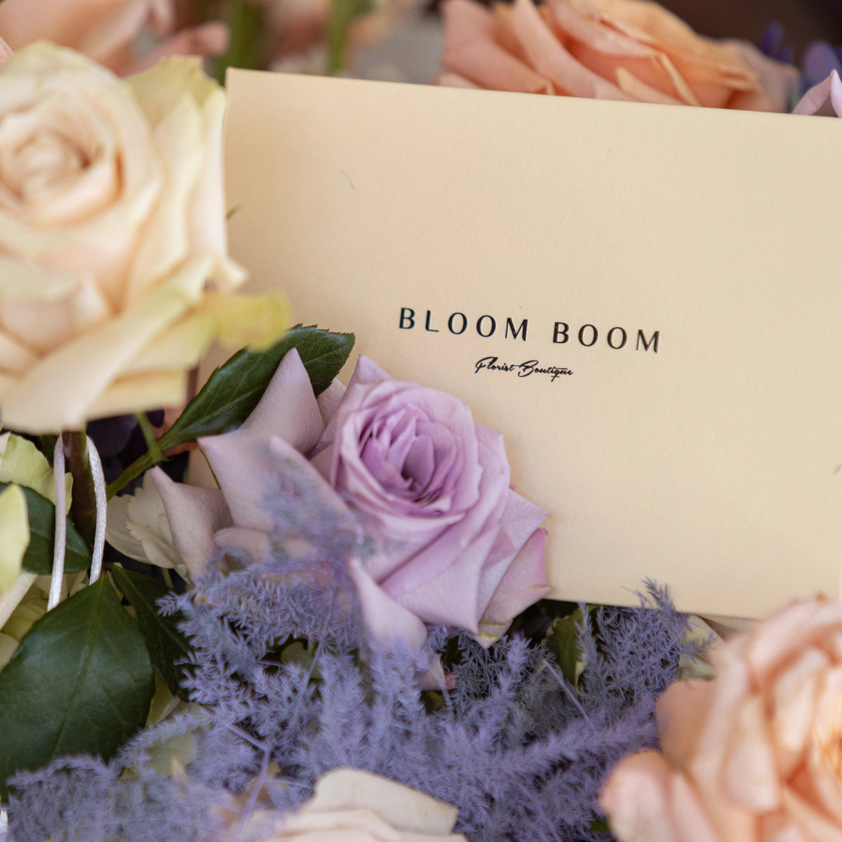 Bloom Boom florist boutique card nestled in pastel flower bouquet with peach roses, purple roses and lavender blooms
