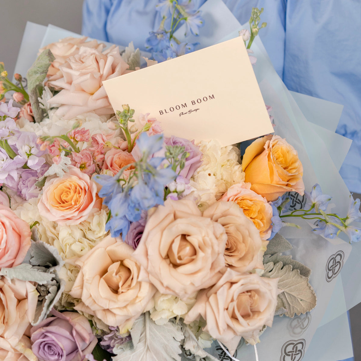 Mixed flower bouquet with peach roses, blue hydrangeas, and Bloom Boom brand card in soft tissue paper wrapping