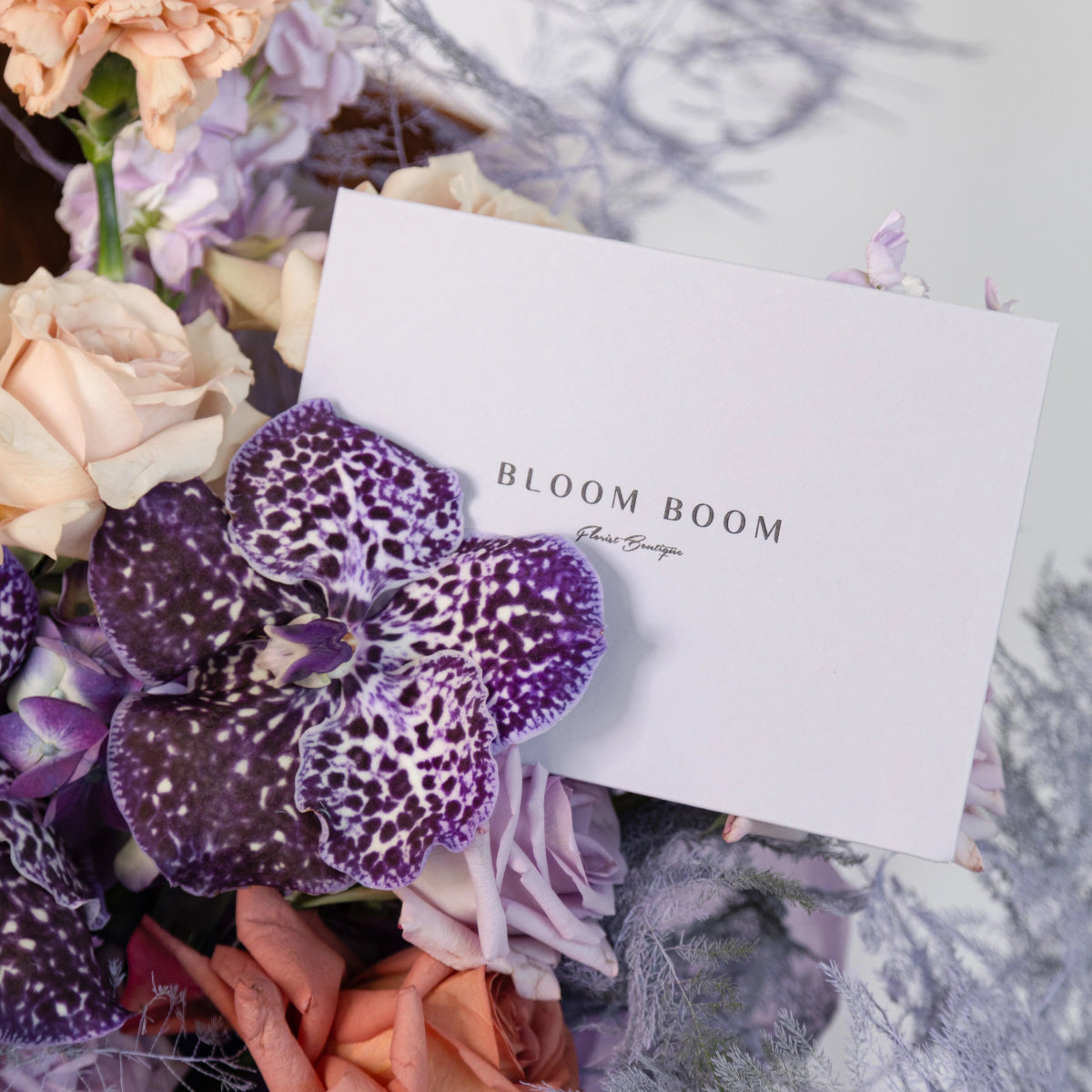 Purple spotted orchid with peach roses and lavender flowers next to white Bloom Boom florist card