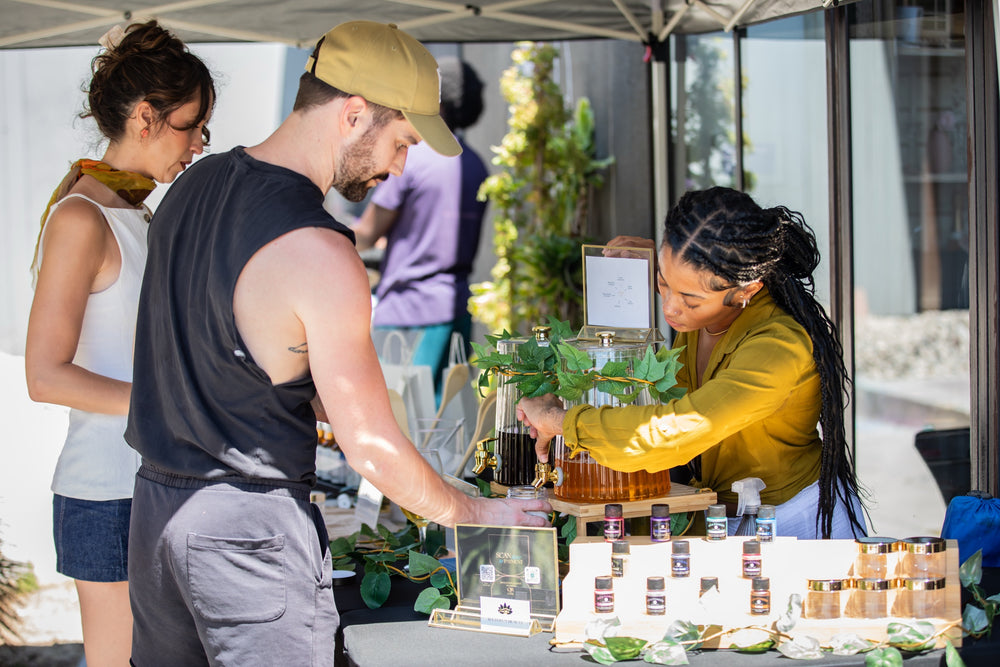 Guests sampling herbal teas and beauty products at a vendor booth under the patio canopy