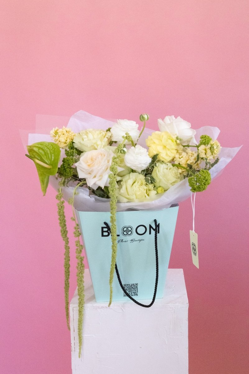 White and yellow roses bouquet in mint green Bloom boutique bag with trailing amaranthus against pink backdrop