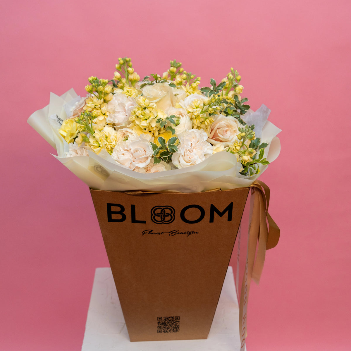 Cream roses and yellow flowers bouquet in brown BLOOM kraft paper bag with white tissue paper on pink background