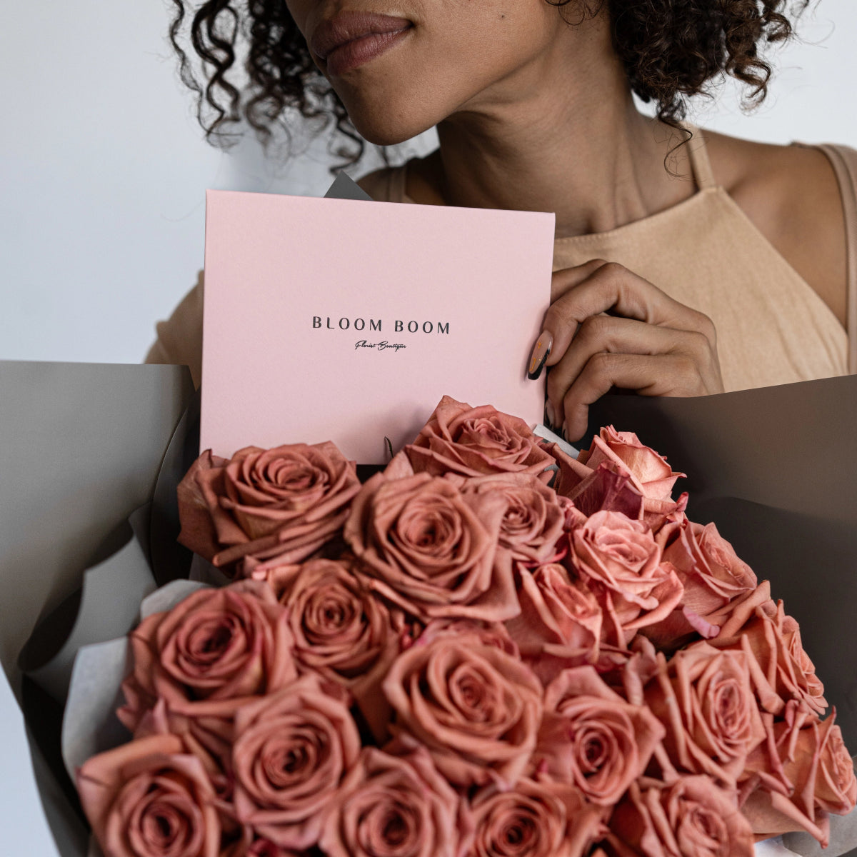Dusty pink roses bouquet in gray box with Bloom Room card held by woman in beige top