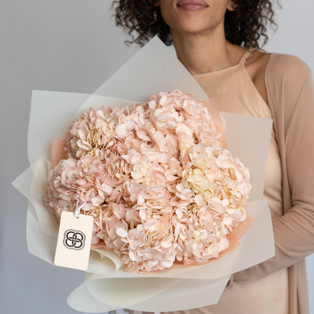 Blush pink hydrangea bouquet wrapped in white paper held by woman in beige dress with brand tag