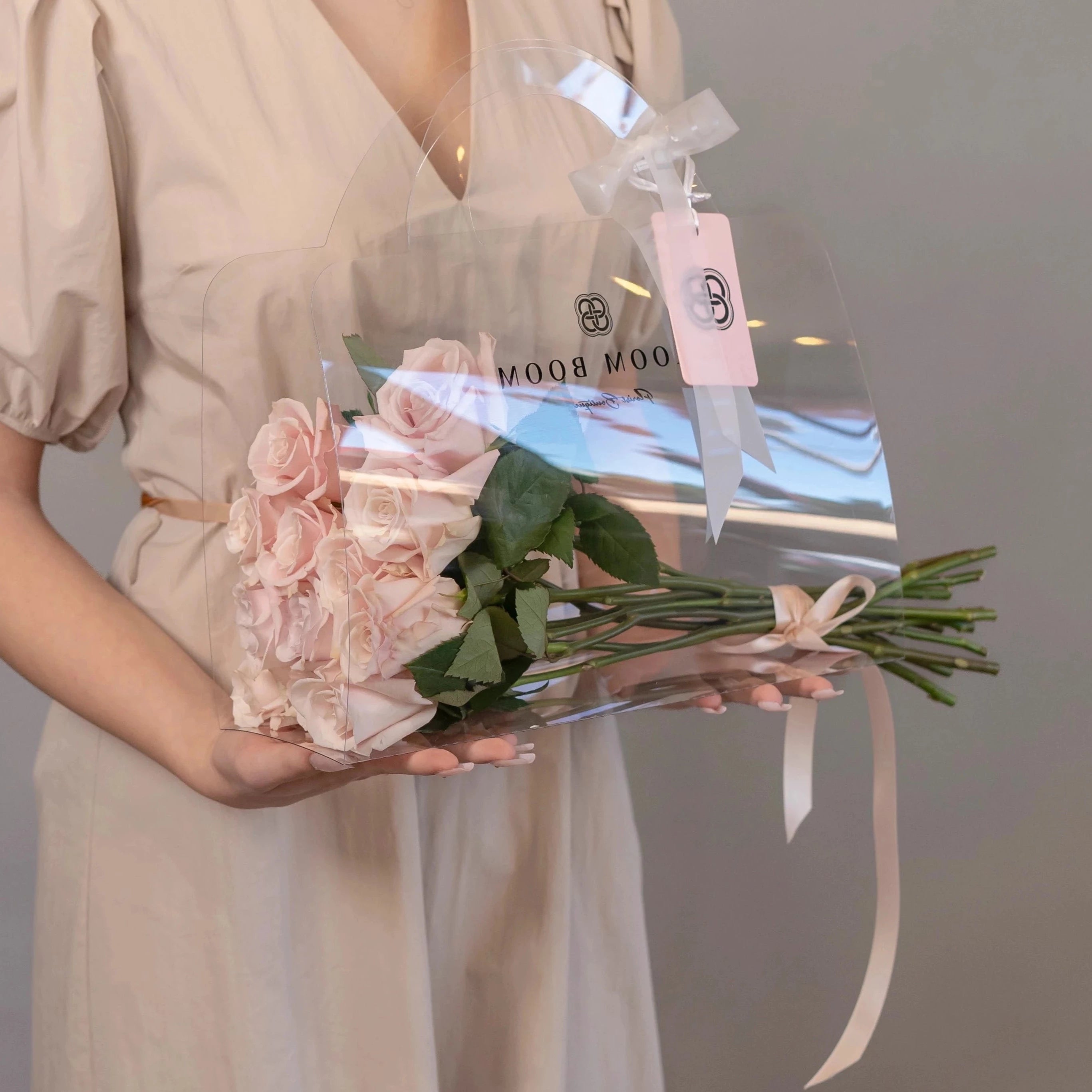 Blush pink rose bouquet with cellophane wrap and Bloom Boom florist tag held by woman in beige dress