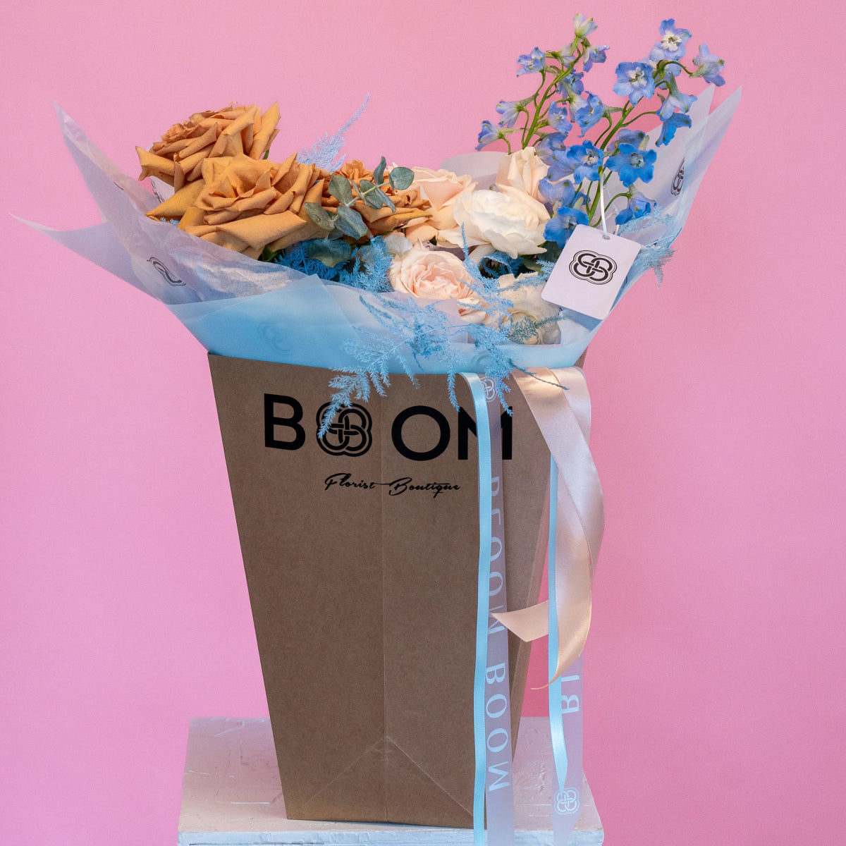 Brown kraft gift bag with mixed flower bouquet featuring orange roses, white roses, and blue delphinium stems