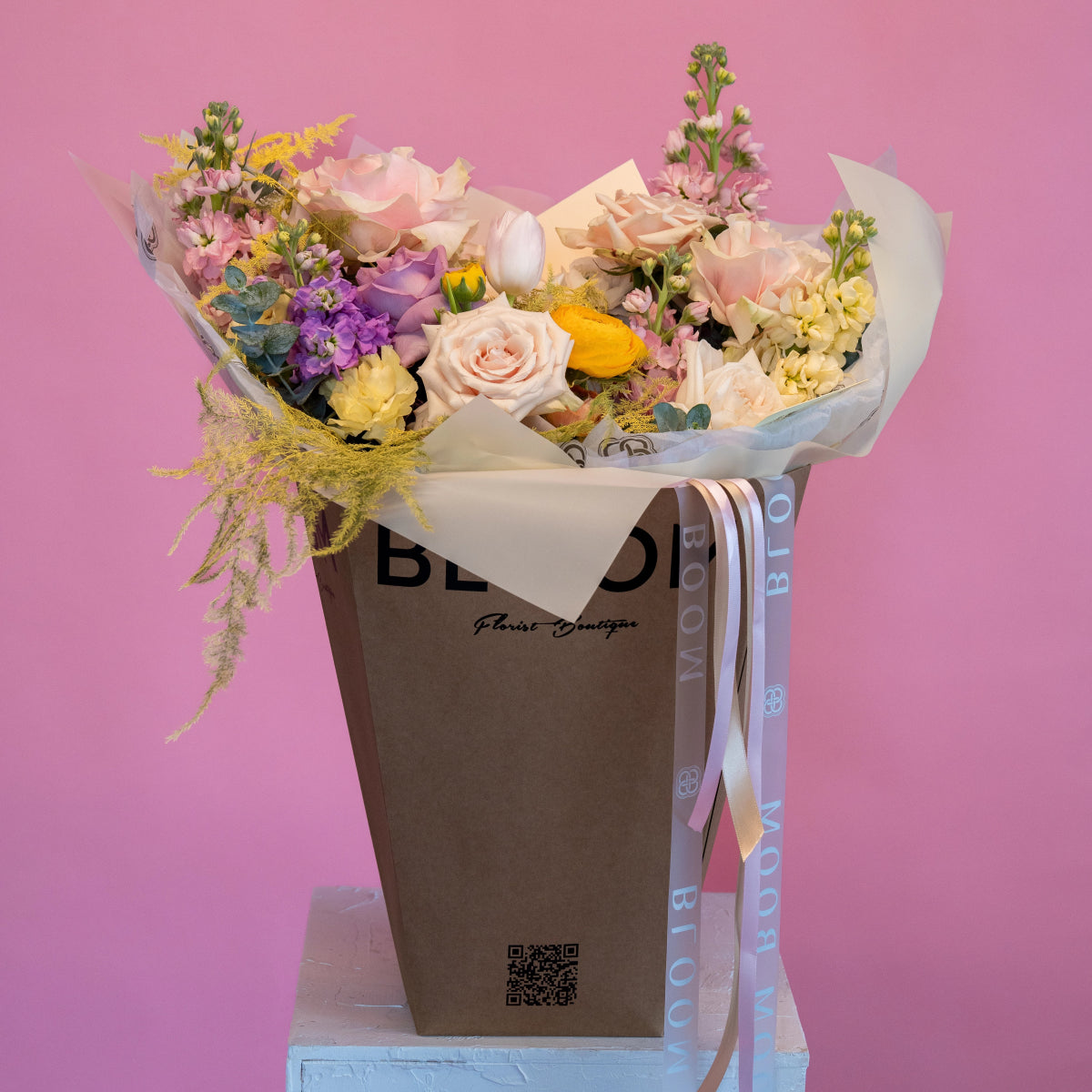 Mixed flower bouquet with pink roses, yellow ranunculus, purple stock in brown paper bag with ribbon on pink background