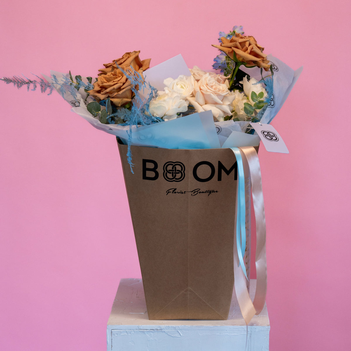 Brown paper gift bag with cream and caramel roses, blue dried flowers, white ranunculus on pink background