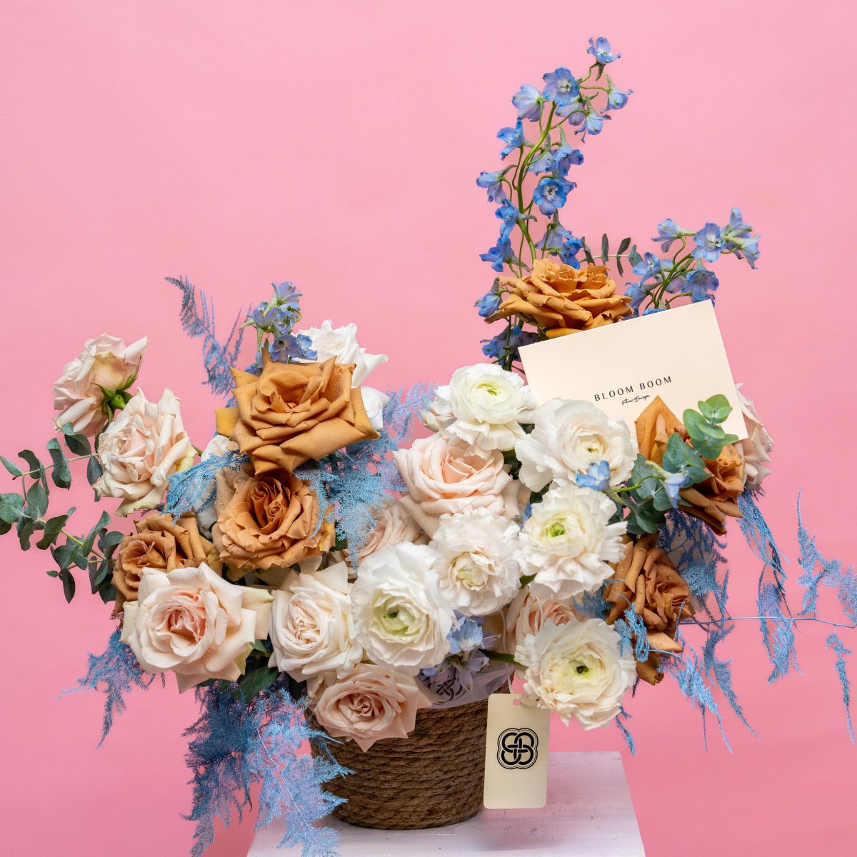 Wicker basket bouquet with peach roses, white ranunculus, blue delphinium on pink background