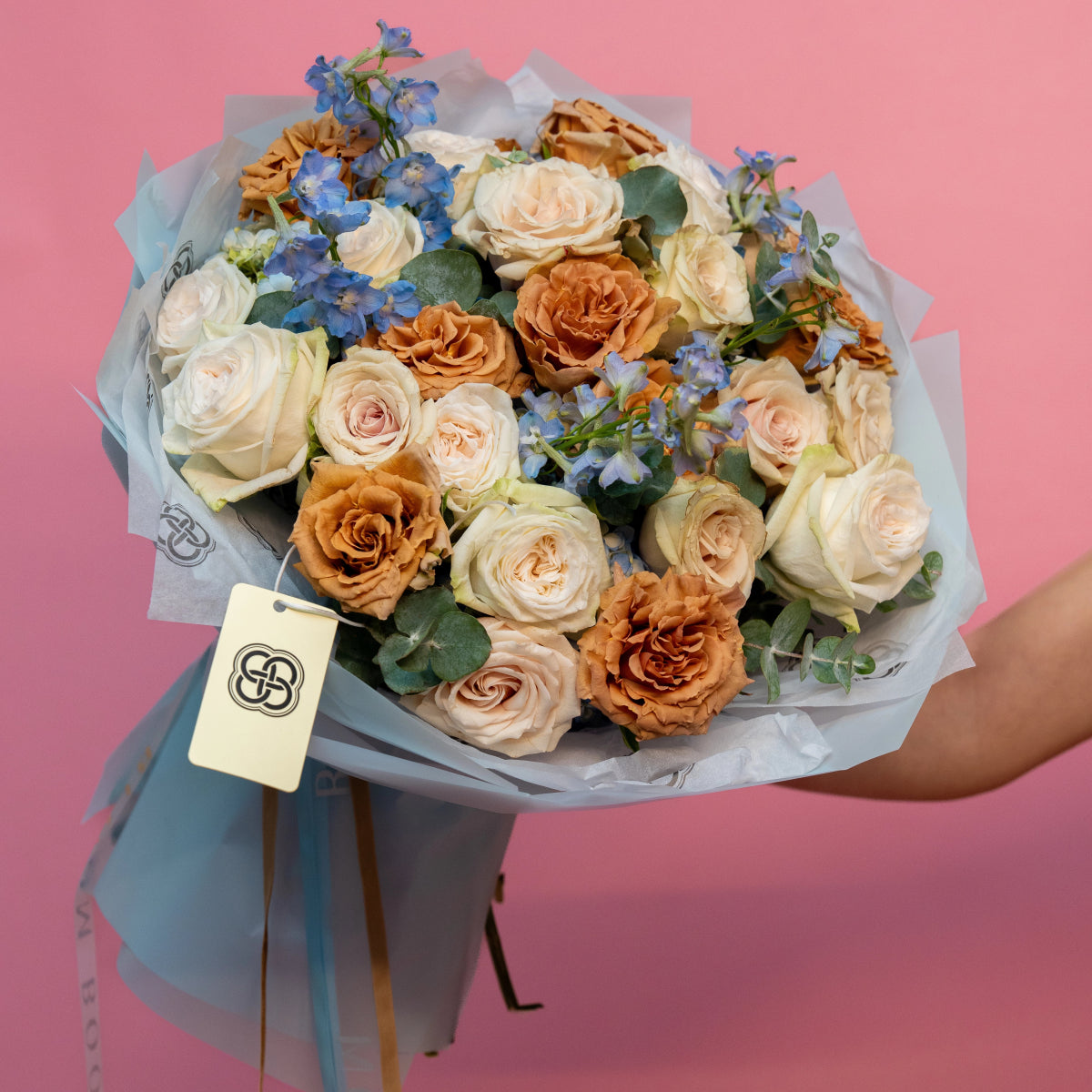 Mixed rose bouquet with cream, peach and bronze roses, blue delphinium flowers wrapped in gray paper with ribbon