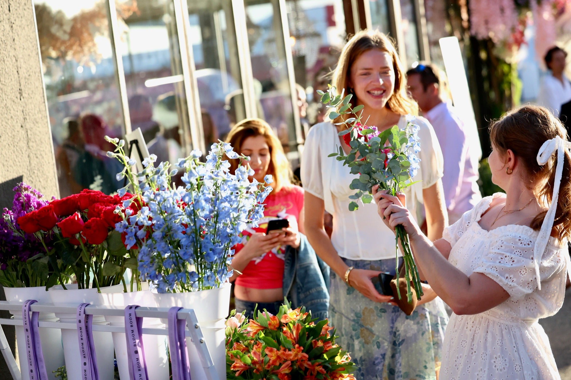 Florist in a white dress handing a bouquet to a guest at a golden-hour sidewalk flower cart with buckets of roses and delphinium