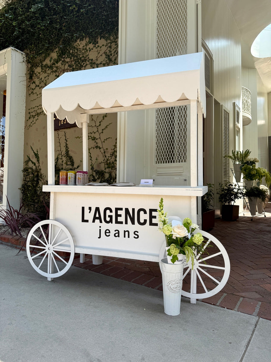 White scalloped-awning flower cart with L’AGENCE lettering and carriage wheels outside an ivy-covered storefront