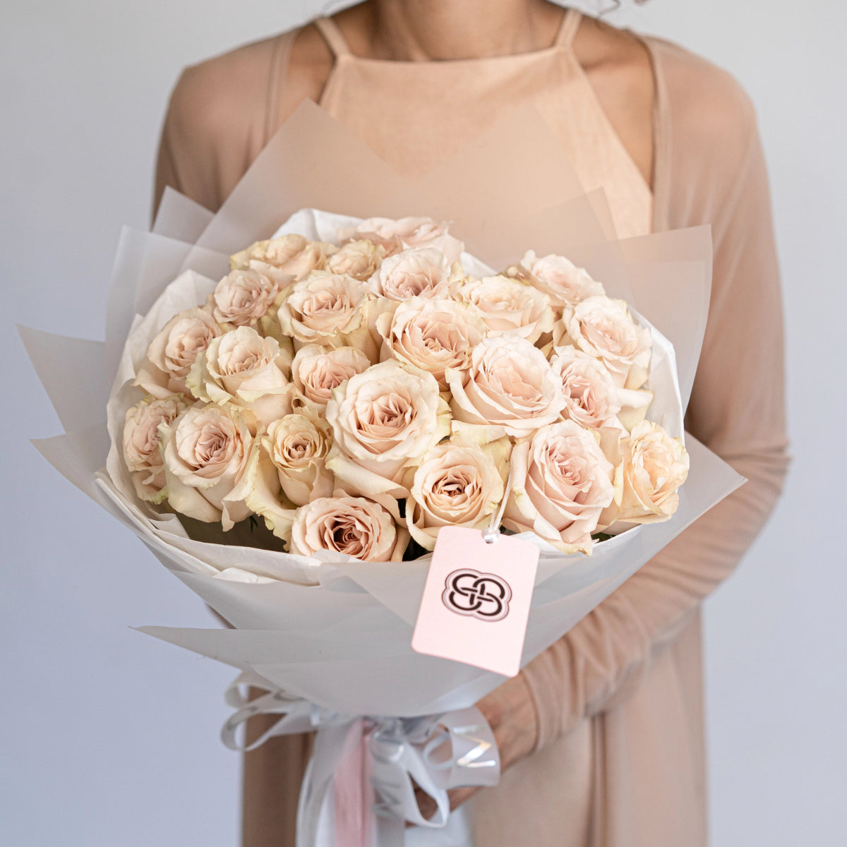 Elegant bouquet of champagne quicksand roses wrapped in white paper with pink ribbon, held by woman in beige dress