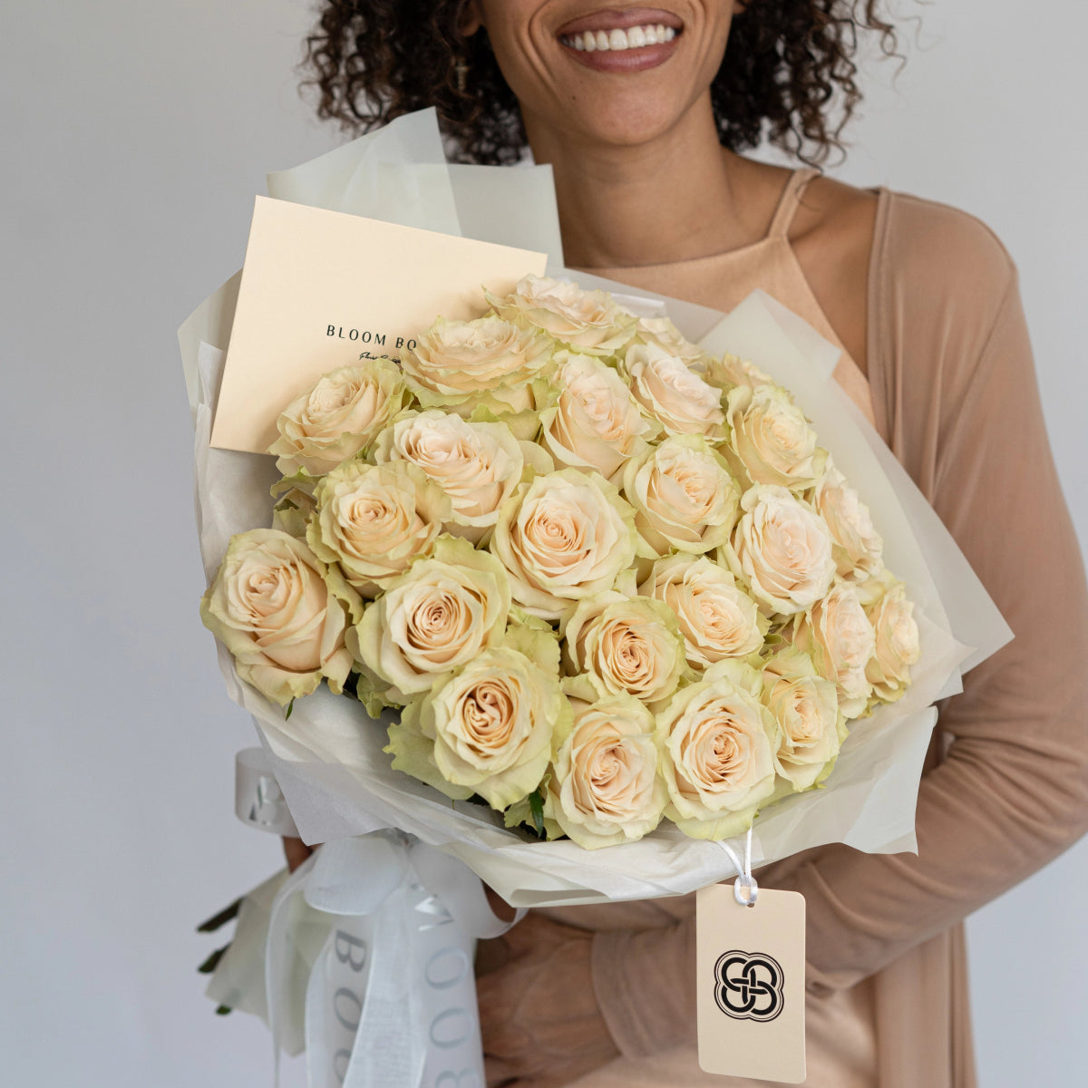 Smiling woman holding large bouquet of champagne-colored roses wrapped in white paper with branded tag