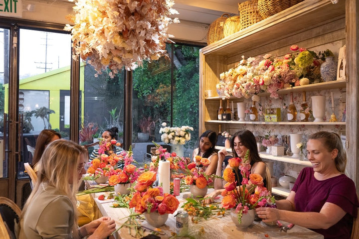 Group of women sitting around a table with floral arrangements in a bright, well-lit room.