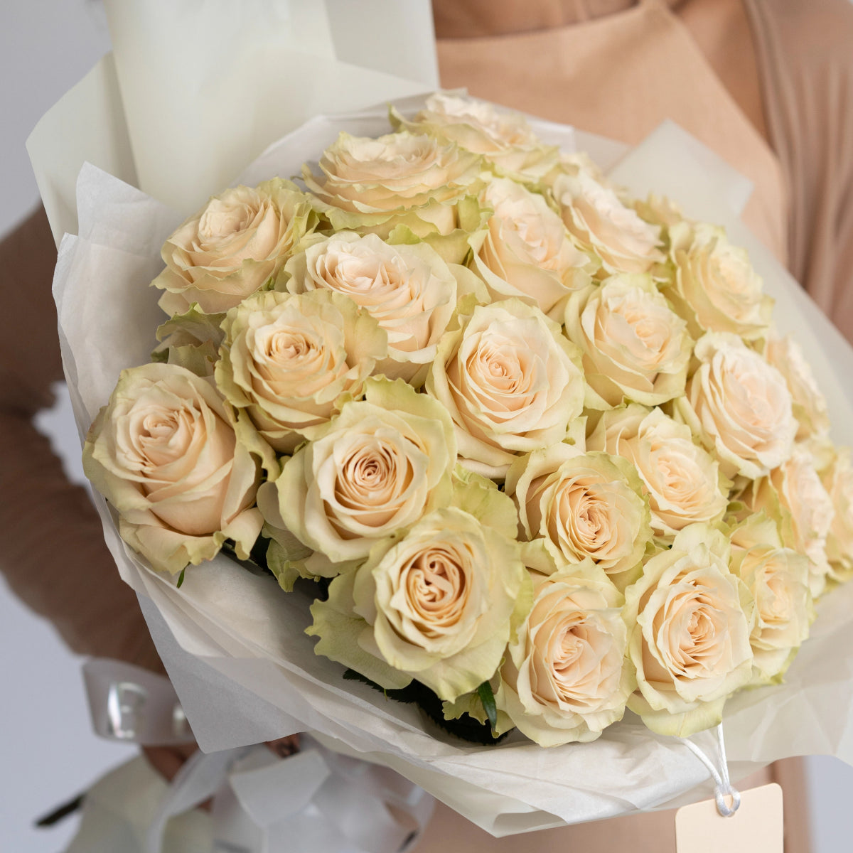 Elegant bouquet of cream and peach roses wrapped in white tissue paper with decorative green accents