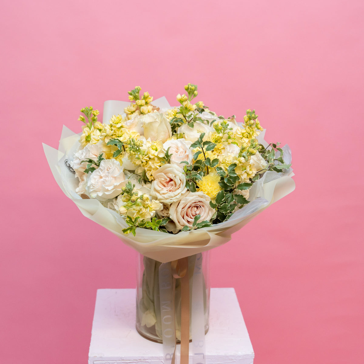 Cream roses and yellow flowers bouquet in glass vase with white paper wrapping against pink background
