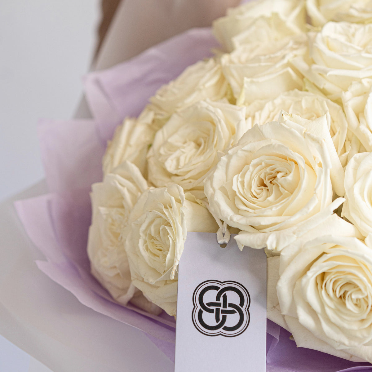 Cream white roses bouquet wrapped in soft lavender tissue paper with elegant black logo ribbon tag