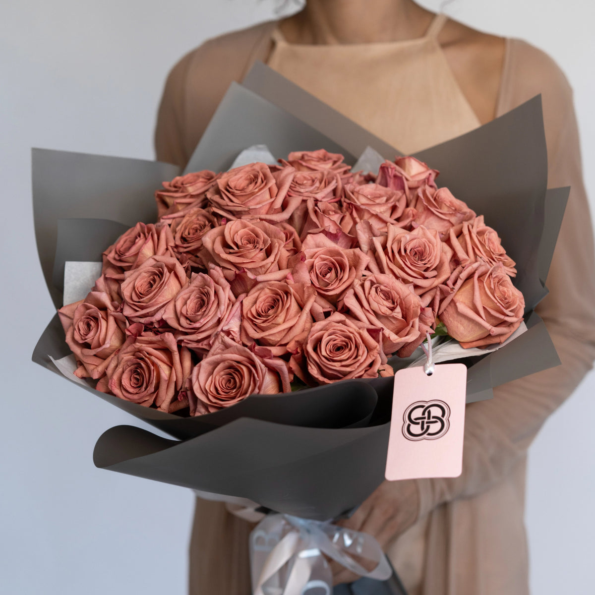 Dusty pink rose bouquet wrapped in charcoal gray paper with brand tag, held by person in beige dress