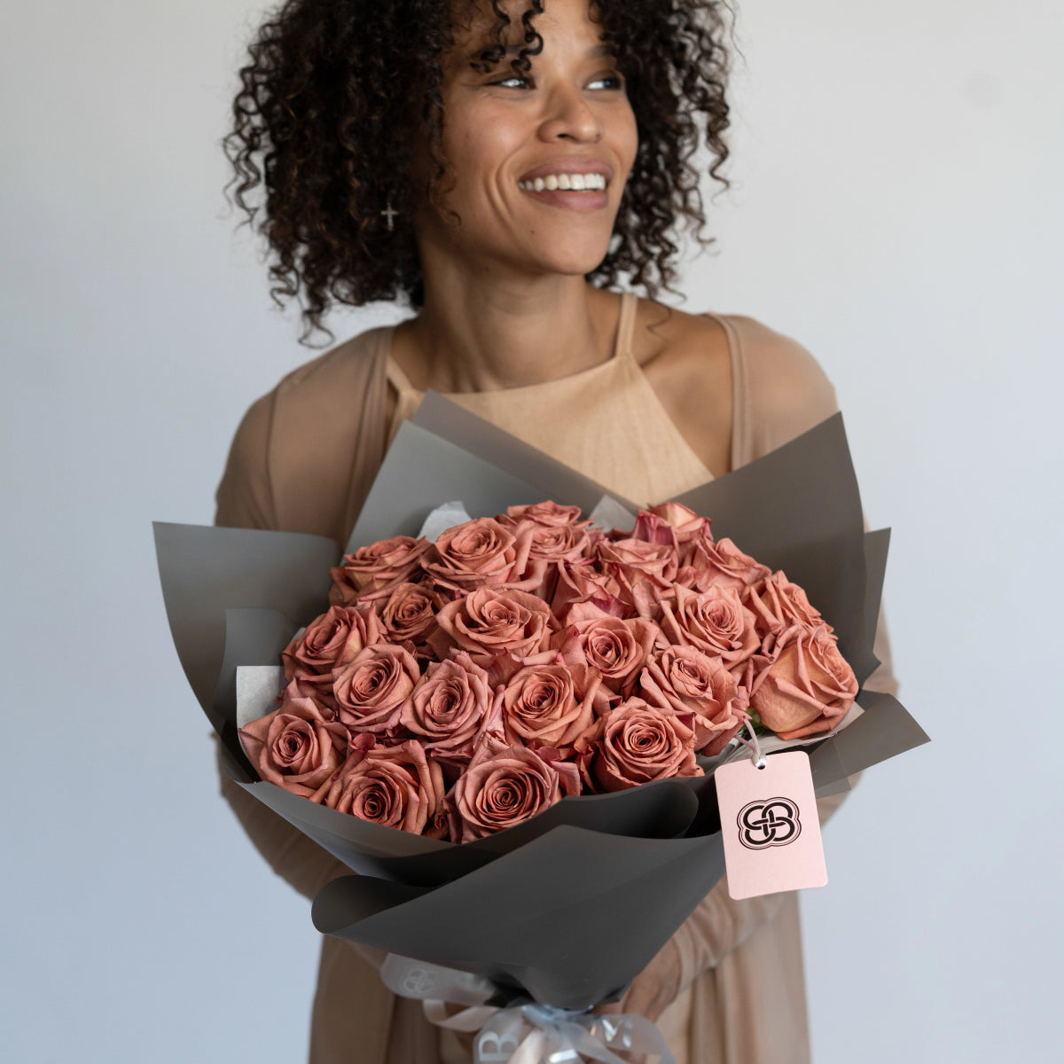 Smiling woman holding large bouquet of dusty rose colored roses wrapped in charcoal gray paper with pink gift tag