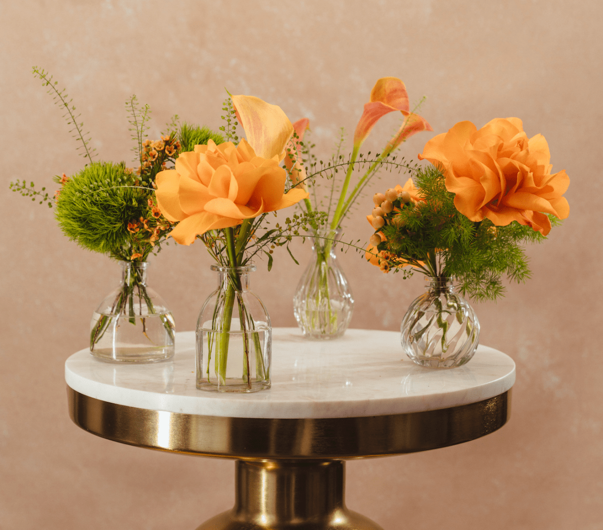 Four glass bud vases with orange flowers and green foliage on white marble table with gold brass base