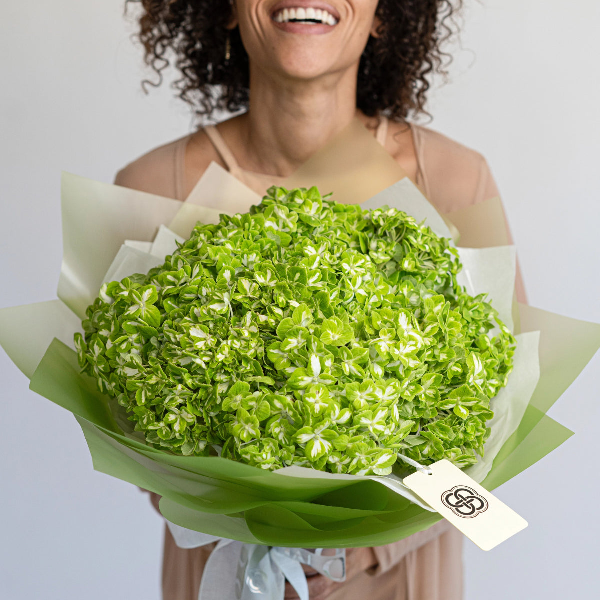 Fresh green hydrangea bouquet wrapped in sage and cream paper with branded tag held by smiling woman