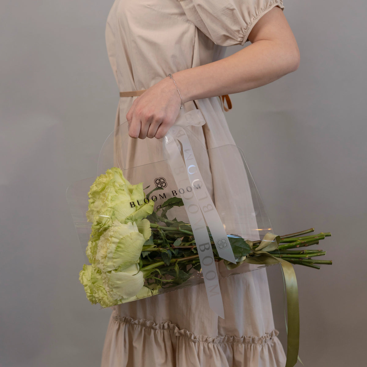 Green roses bouquet with clear cellophane wrapping and white Bloom Box ribbon held by woman in beige dress