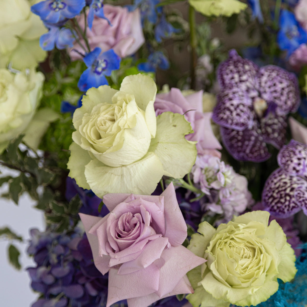 Mixed flower bouquet with cream and purple roses, blue delphinium, and textured purple blooms in garden arrangement