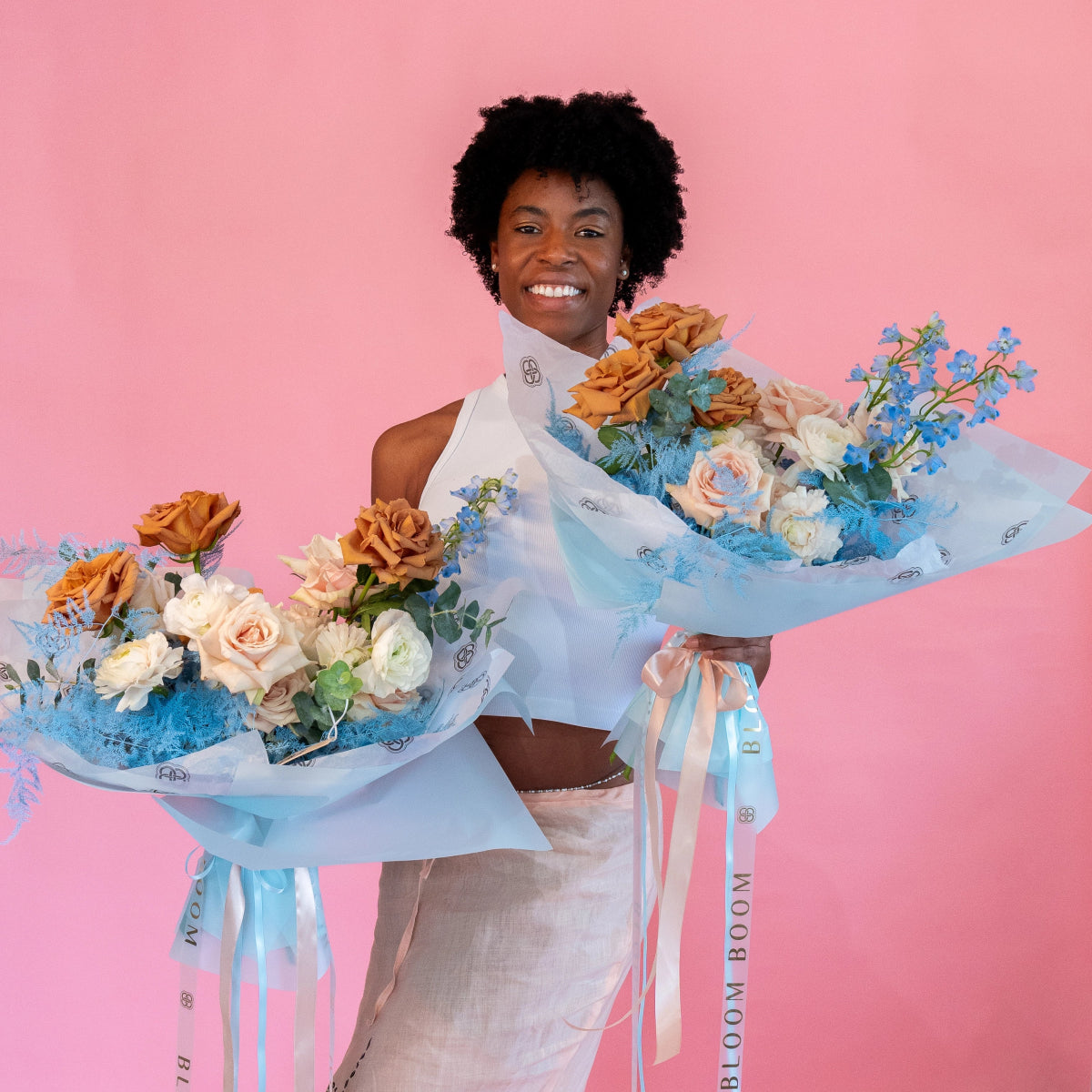 Woman holding two mixed flower bouquets with orange roses, pink ranunculus, and blue accents in light blue wrapping