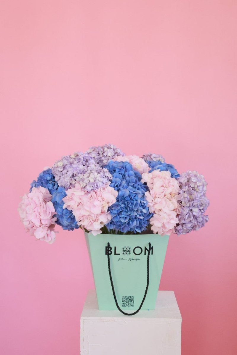 Mixed hydrangea bouquet in mint green Bloom bag with pink, blue, and purple flowers on white pedestal