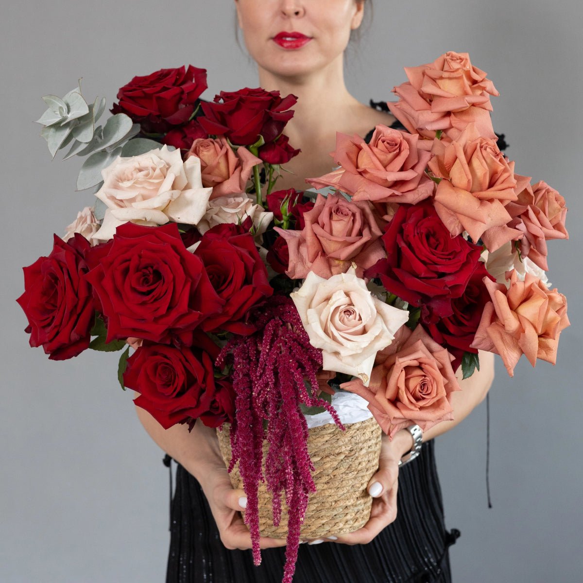 Mixed rose bouquet in woven basket with red, coral, and white roses plus burgundy amaranthus and eucalyptus