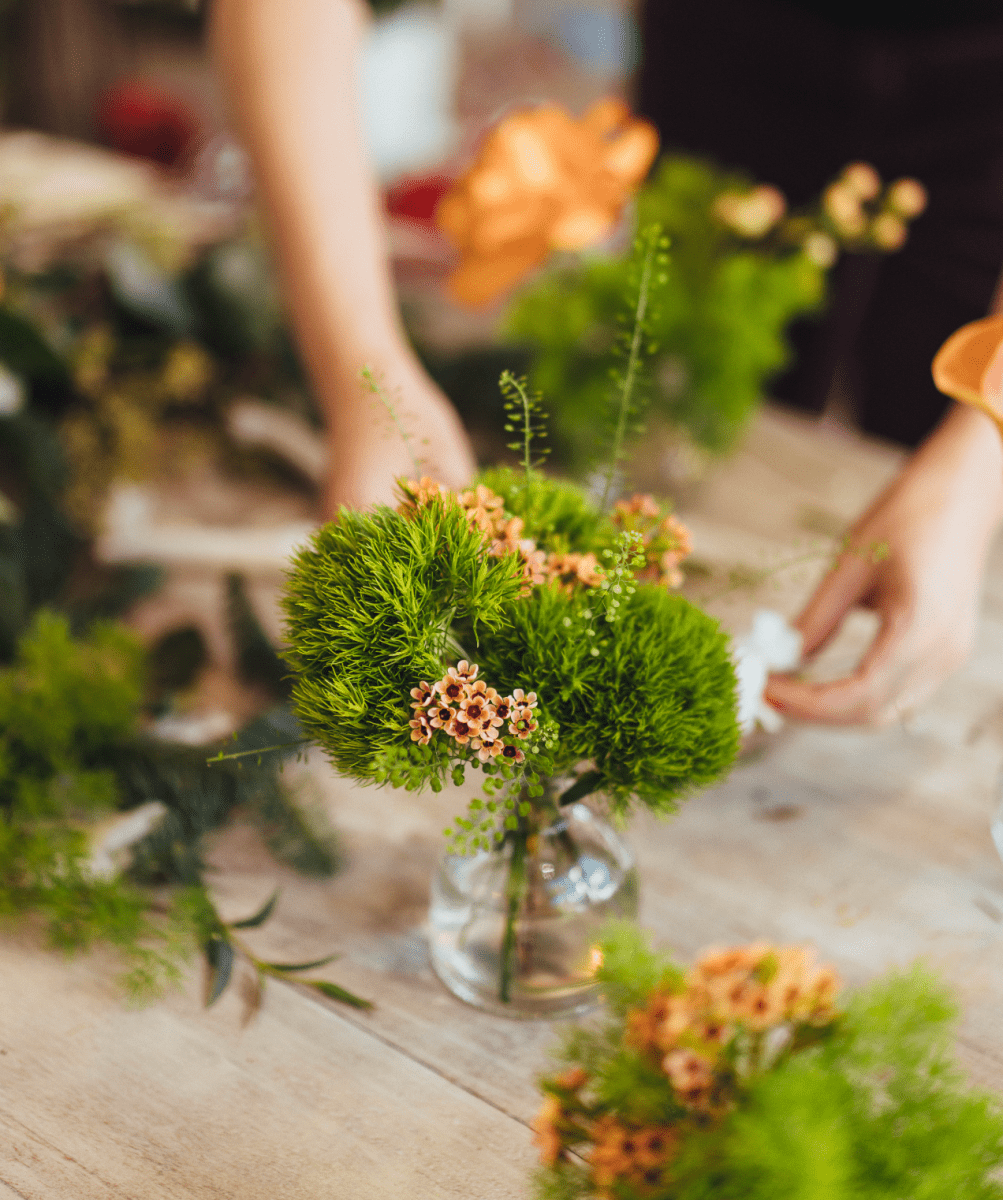 Green moss ball centerpiece with small pink flowers in clear glass vase on wooden table during floral workshop