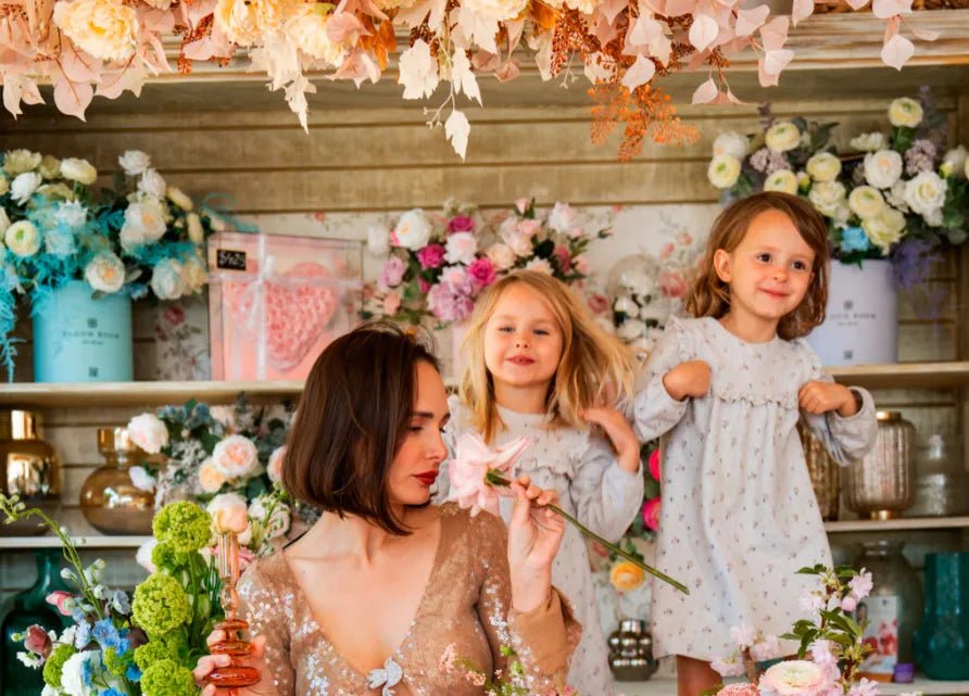Mother in gold sequin dress with two young girls in white floral dresses surrounded by colorful spring flowers