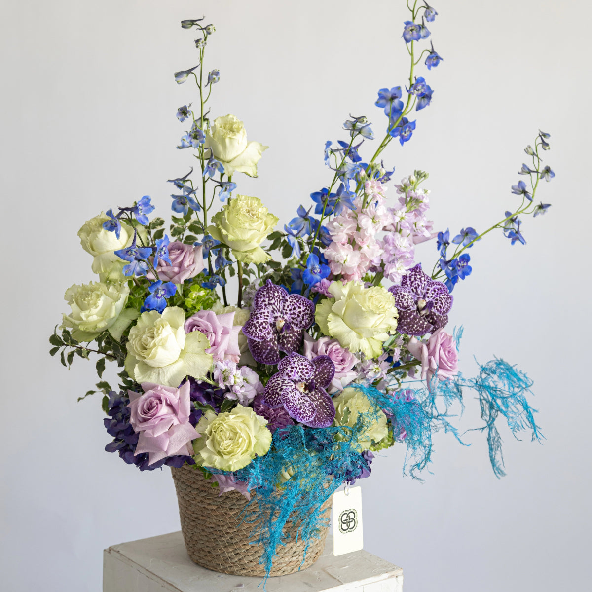 Wicker basket arrangement with white roses, purple orchids, blue delphiniums, and turquoise decorative moss
