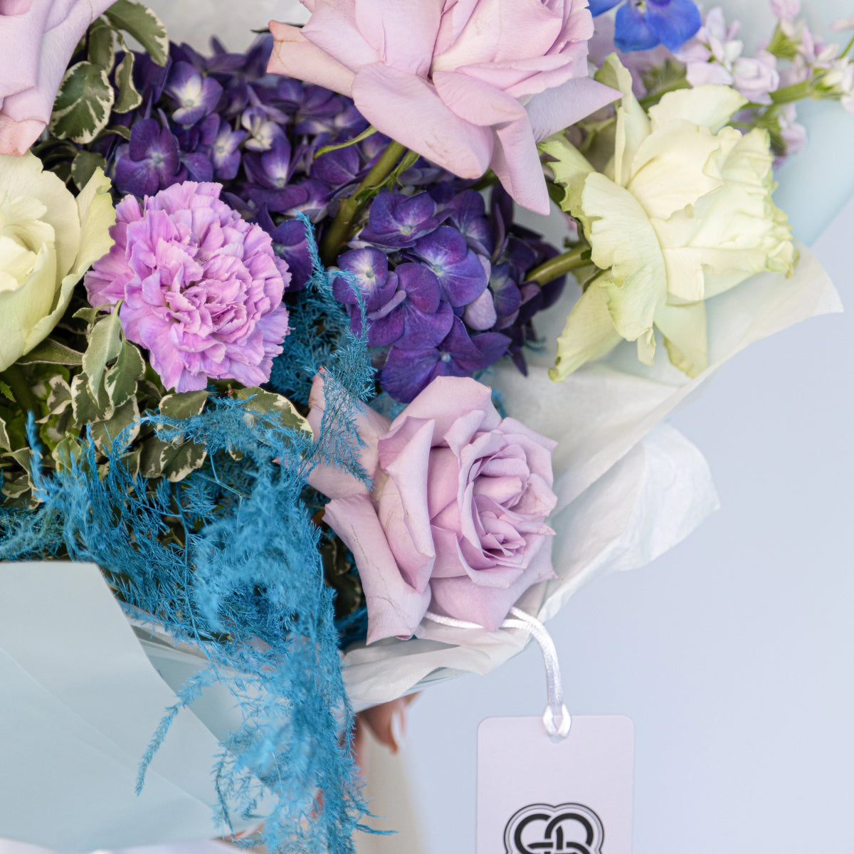 Mixed flower bouquet with purple roses, lavender carnations, blue hydrangea and teal accent foliage in white paper