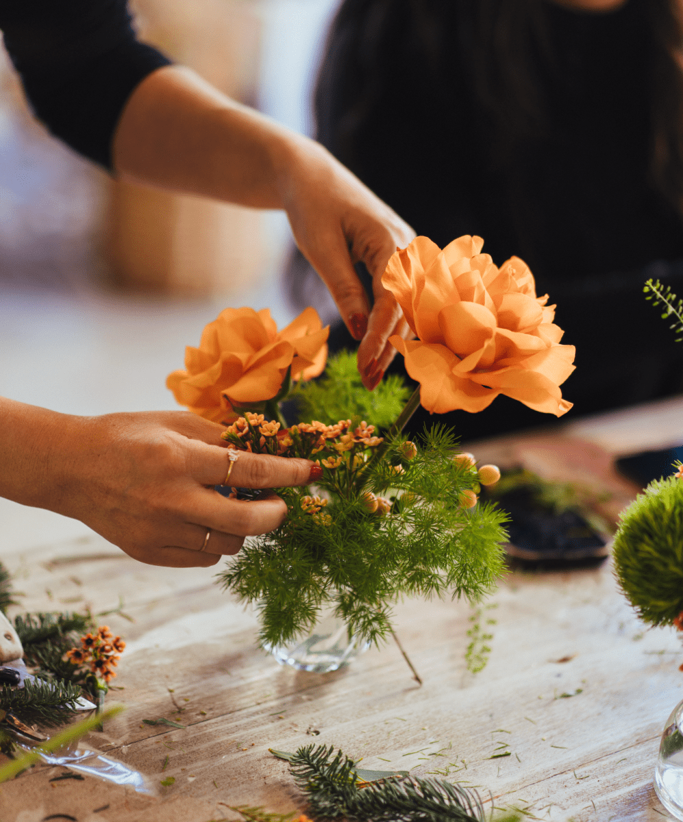 Hands arranging orange carnation flowers with green ferns in glass vase for floral arrangement workshop