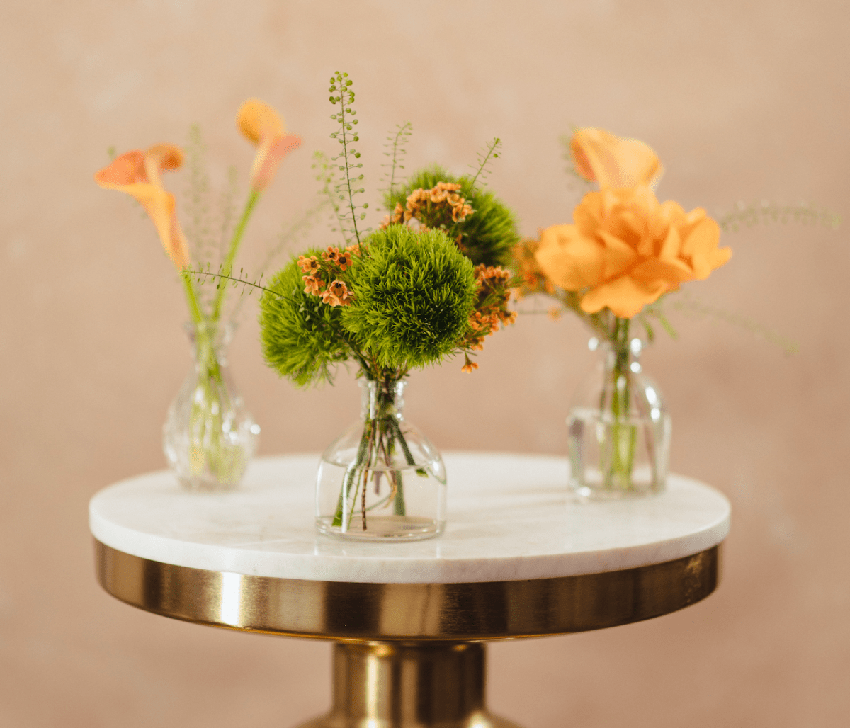 Three clear glass vases with orange flowers and green moss balls on white marble table with gold brass base