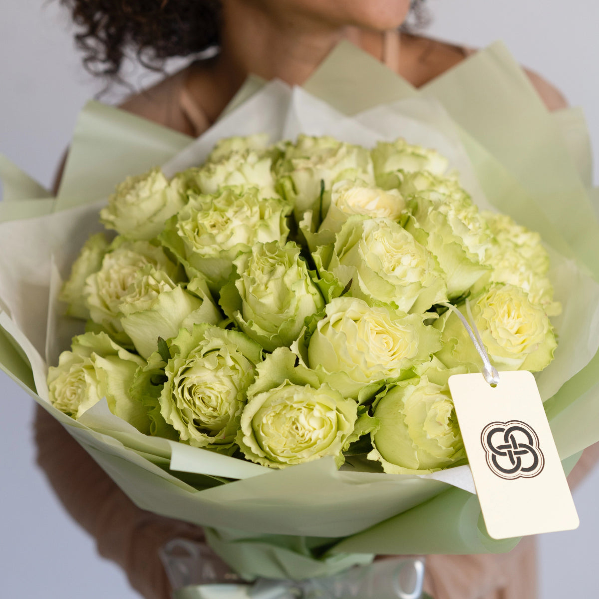 Fresh pale green rose bouquet wrapped in light green paper with white gift tag held by woman