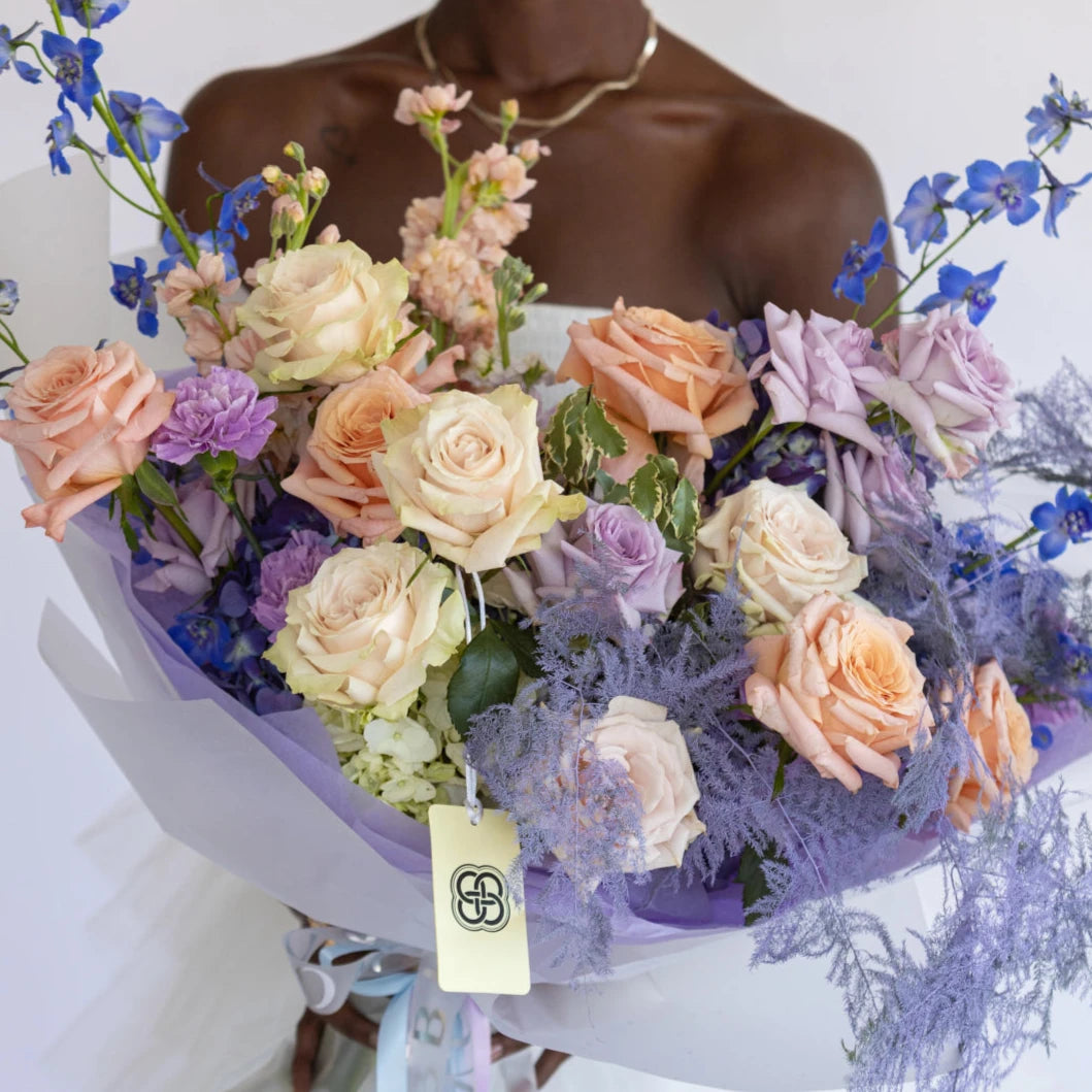 Mixed pastel bouquet with peach, cream and lavender roses, blue delphinium, purple dusty miller in white wrap