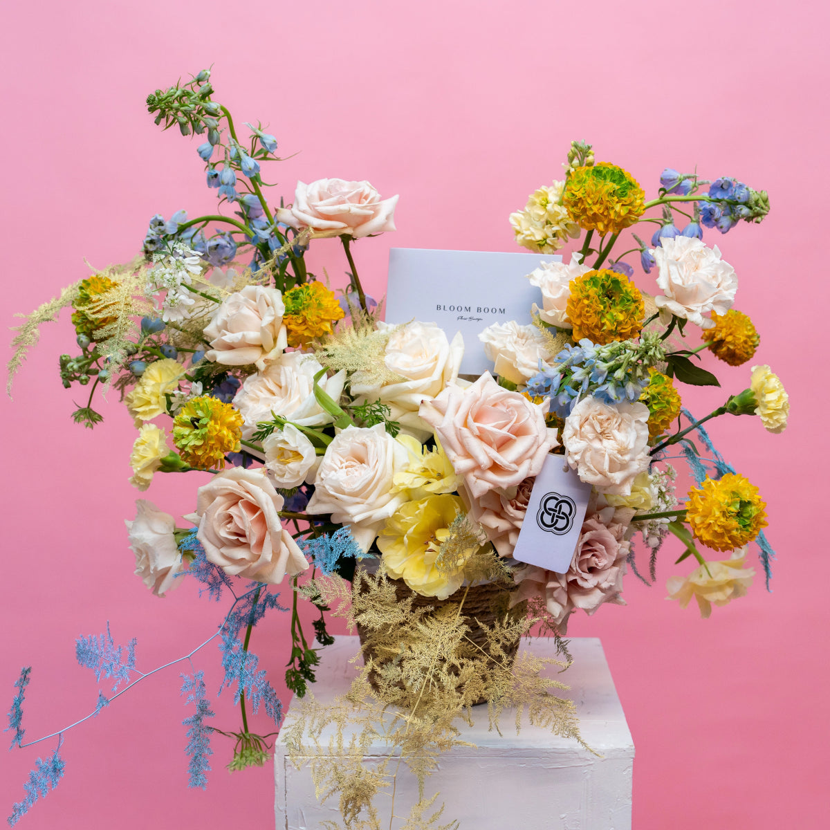 Pastel rose bouquet with yellow marigolds and blue delphinium in clear vase against pink background