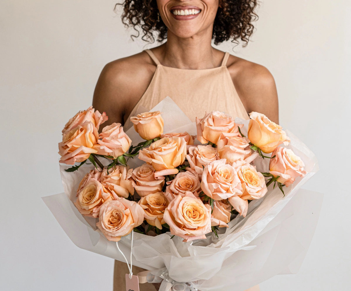 Large peach coral rose bouquet wrapped in white paper held by smiling woman in nude halter top