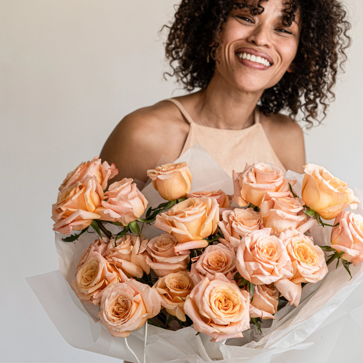 Smiling woman holding large bouquet of peach and coral roses wrapped in white tissue paper against neutral background