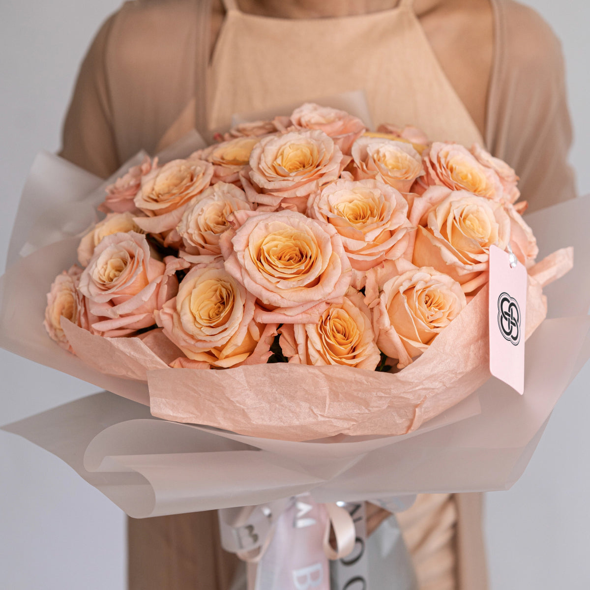 Peach and cream colored roses wrapped in soft pink tissue paper with satin ribbon, held by person in beige outfit